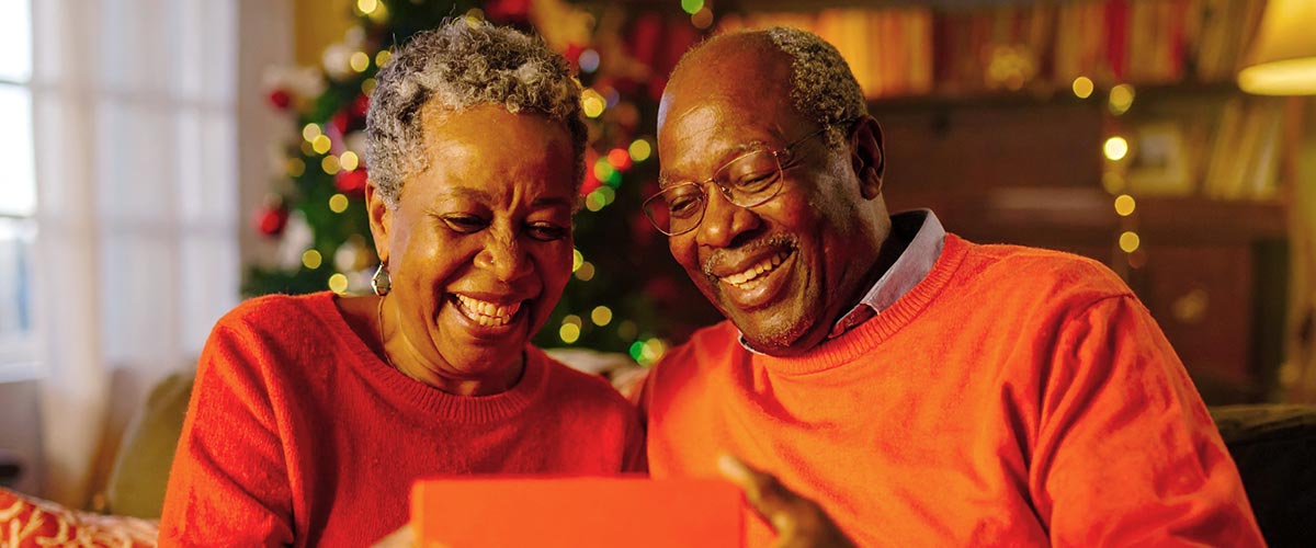 Senior couple smiling with a gift box, showcasing a Forged Flare Christmas ornament for grandparents, surrounded by festive holiday decorations.