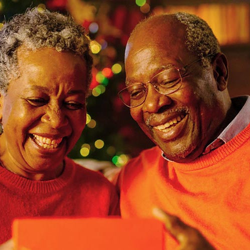 Senior couple smiling with a gift box, showcasing a Forged Flare Christmas ornament for grandparents, surrounded by festive holiday decorations.