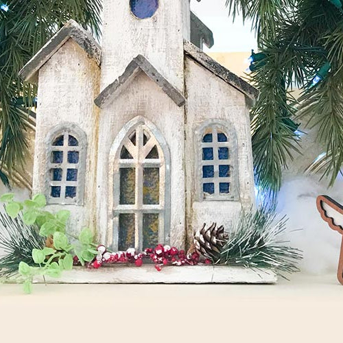 Handcrafted Christmas ornament display with Forged Flare® Mother’s Angels® angels, a small church model, and festive greenery for a cozy holiday setup.