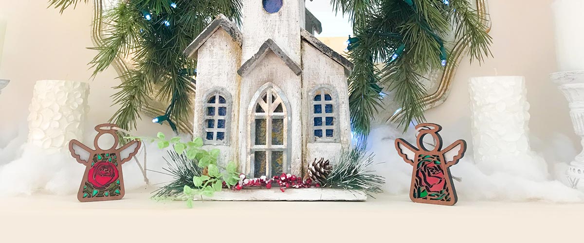 Handcrafted Christmas ornament display with Forged Flare® Mother’s Angels® angels, a small church model, and festive greenery for a cozy holiday setup.