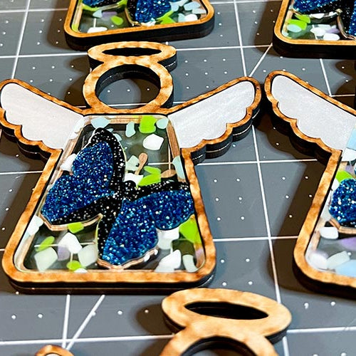 Handcrafted angel ornaments by Forged Flare® in Texas. Multiple angel figurines with blue glittery butterfly wings on a grid surface.