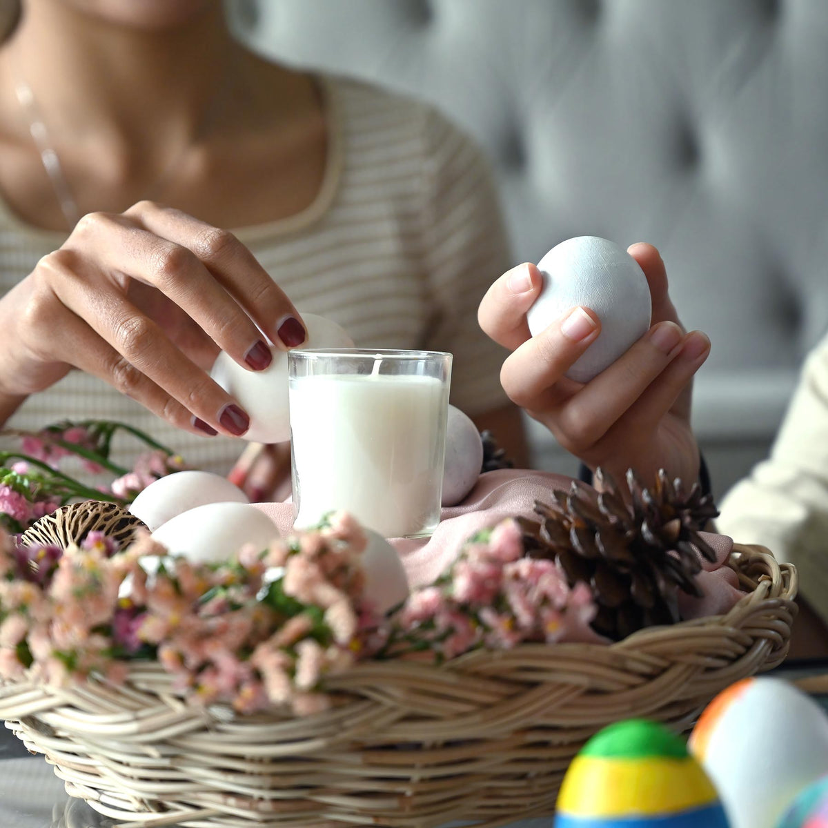 A person preparing an Easter basket with delicate Easter decorations, holding a white egg near a candle, with a variety of Easter ornaments and colorful eggs in the background, offering inspiration for Easter basket ideas.