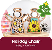 Two Forged Flare® Mother's Angels® 3.5" holiday figurines—one with a daisy, one with a sunflower—are displayed. A green "Save 10%" badge is shown, with "Holiday Cheer: Daisy + Sunflower" below. A pink Christmas tree appears blurred in the background.