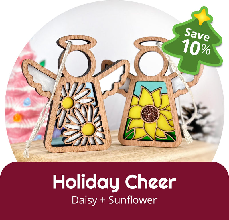Two Forged Flare® Mother's Angels® 3.5" holiday figurines—one with a daisy, one with a sunflower—are displayed. A green "Save 10%" badge is shown, with "Holiday Cheer: Daisy + Sunflower" below. A pink Christmas tree appears blurred in the background.