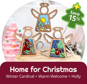 The Forged Flare® Home for Christmas 3-Piece Bundle features 3.5" Mother's Angels® figurines—Winter Cardinal, Warm Welcome, and Holly. “Home for Christmas” text and a “Save 15%” green badge; a pink Christmas tree appears blurred in the background.