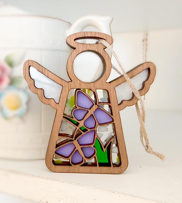 A wooden angel ornament with stained-glass-style purple butterflies, green leaves, and a white hanging loop, shown on a light surface with a soft blurred background.