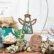 The Snowdrop Ornament, a 3.5" angel figurine from the Mother's Angels® collection by Forged Flare®, hangs gracefully on a stand. Nearby are ribbon curls, a cupcake with sprinkles, and a wrapped gift with a floral design, along with a tag that reads "Happy Birthday." The background showcases part of a kitchen. This charming and festive scene is ideal for birthday gifts for women.