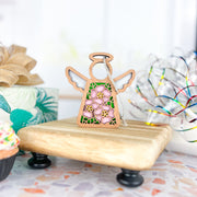 The Forged Flare® Pink Evening Primrose Ornament, a 3.5" Mother's Angels® angel figurine with floral patterns, stands on a riser next to a ribboned gift, colorful cupcake, and shiny ribbons.