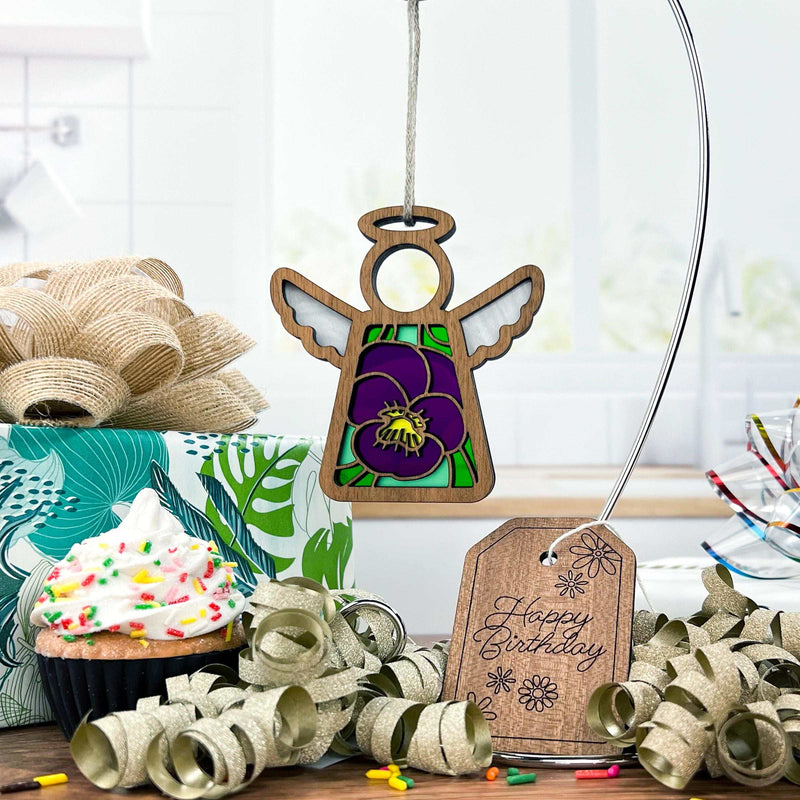 A Forged Flare® Violet Ornament | 3.5" Angel Figurine from Mother's Angels® hangs above a sprinkled cupcake, wrapped gift, curled ribbon, and a “Happy Birthday” tag on a table in a bright kitchen.