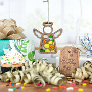The Forged Flare® Daffodil Ornament (3.5" H Mother's Angels® angel figurine) sits by a "Happy Birthday" tag, ribbon curls, cupcake, gift box, and confetti on a festive table—an ideal birth flower gift.