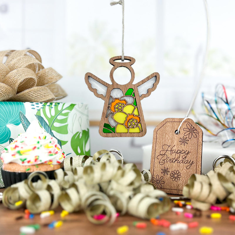 The Forged Flare® Daffodil Ornament (3.5" H Mother's Angels® angel figurine) sits by a "Happy Birthday" tag, ribbon curls, cupcake, gift box, and confetti on a festive table—an ideal birth flower gift.