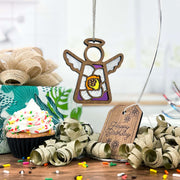 A Forged Flare® Mother's Angels® Jonquil Ornament (3.5" angel figurine) hangs over a sprinkle-topped cupcake, beside a wrapped gift, gold ribbon curls, and a "Happy Birthday" tag. A white jonquil flower—the March birth flower—completes the scene.