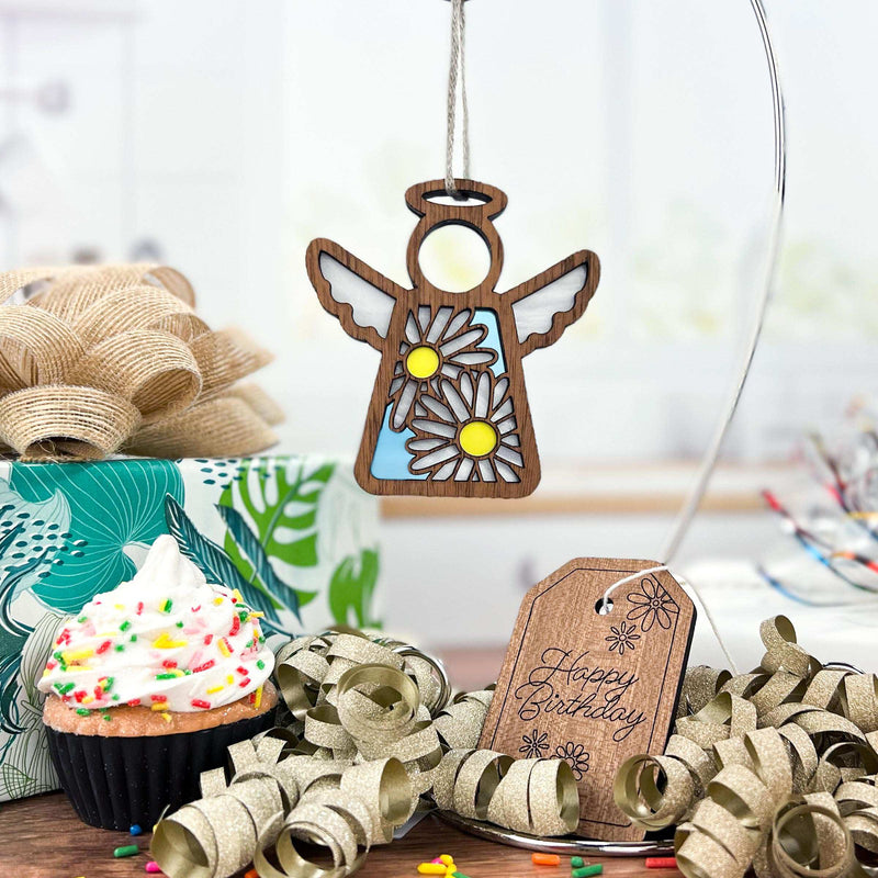 A Forged Flare® Mother’s Angels® 3.5" Daisy Ornament angel figurine hangs near a sprinkled cupcake, gold ribbon, floral gift box, and wooden “Happy Birthday” tag on a bright, festive table.