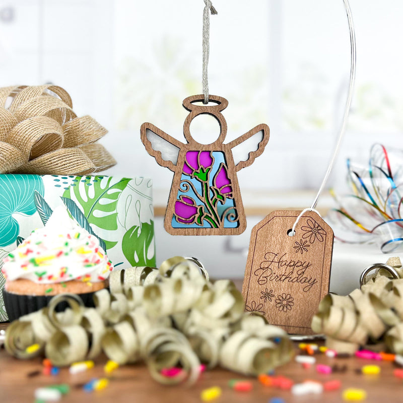A Forged Flare® Sweet Pea Ornament, a 3.5" Mother's Angels® angel figurine with a stained-glass center, hangs with birthday decor—wrapped gift, curled ribbons, Happy Birthday tag, and sweet pea flower accents for a special touch.