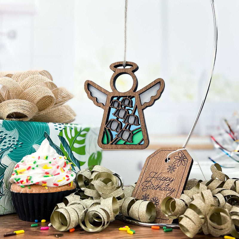 A Forged Flare® Lily of the Valley Ornament—Mother's Angels® 3.5" angel—hangs before a sprinkled cupcake birthday gift with gold ribbon, a “Happy Birthday” tag, lily of the valley accents, wrapping paper, and festive touches.