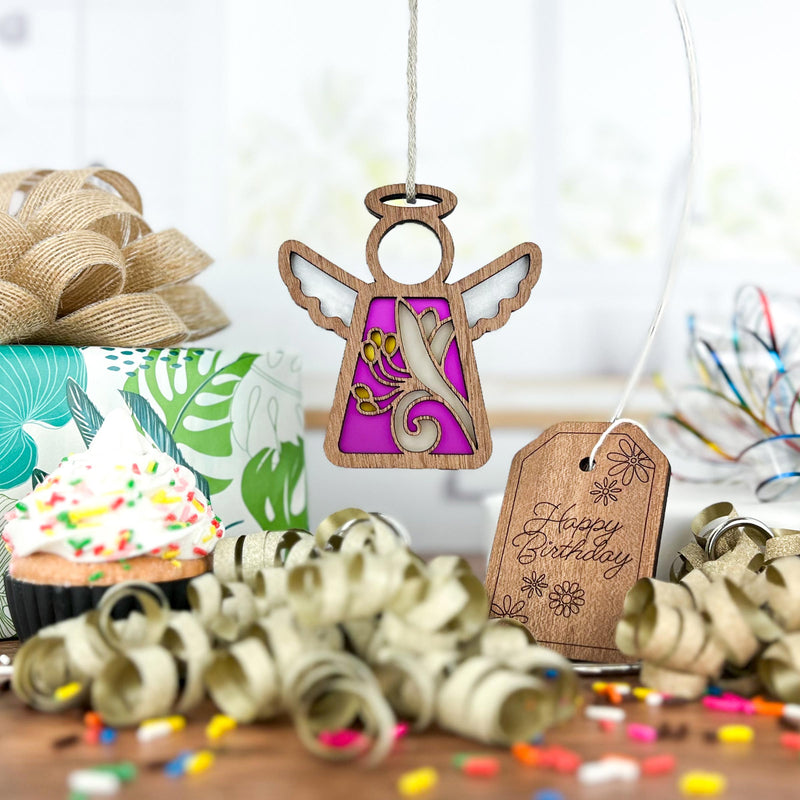 A Forged Flare® Honeysuckle Ornament (3.5" angel figurine from Mother's Angels®), featuring purple and gold, hangs above a sprinkle-topped cupcake. A birth flower tag, curling ribbon, confetti, and gift box add birthday cheer.