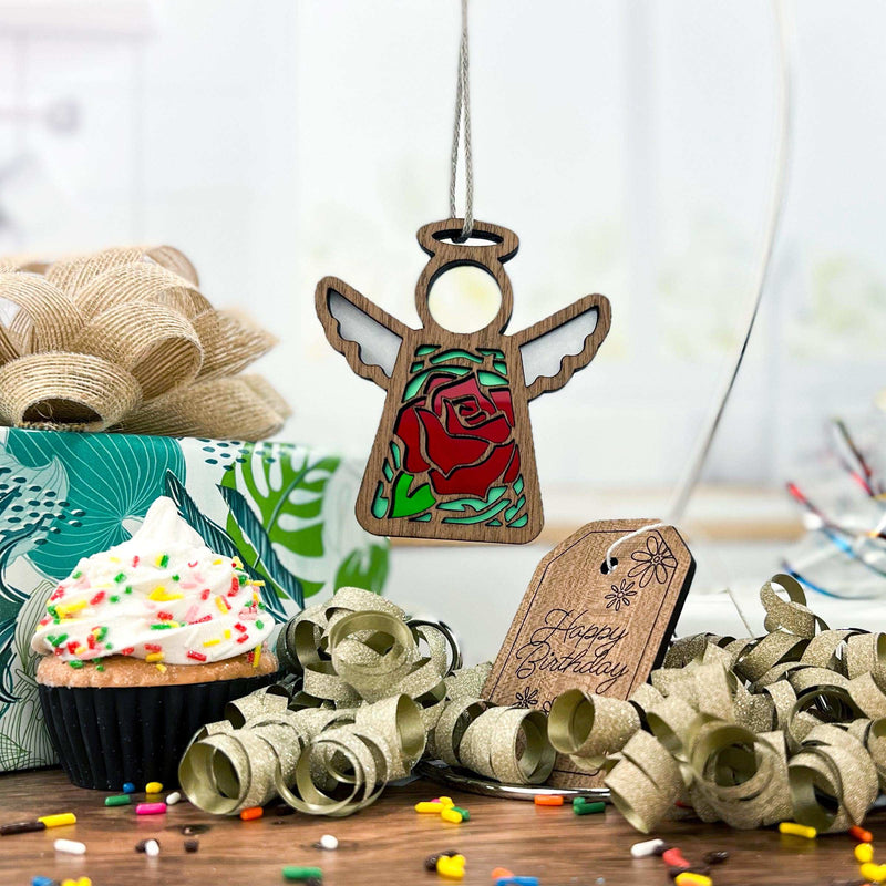 Display a Forged Flare® Mother’s Angels® Red Rose Ornament with Imperfections, a 3.5" handcrafted angel figurine, above a festive birthday scene featuring a wrapped gift, sprinkle cupcake, gold ribbon curls, and confetti on wood.