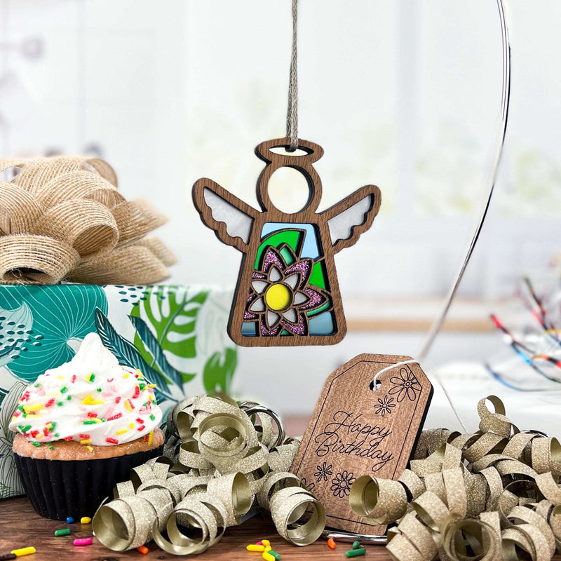 A Forged Flare® Water Lily Ornament | 3.5" (H) Angel Figurine from the Mother's Angels® collection hangs above a table with a sprinkle-topped cupcake, gold ribbon curls, a "Happy Birthday" tag, burlap bow, and birth flowers.