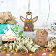 The Forged Flare® Mother's Angels® Aster Ornament—a 3.5" wooden angel with purple aster flower details—hangs by a “Happy Birthday” tag, surrounded by a gift box, cupcake with sprinkles, curly ribbons, and a burlap bow.