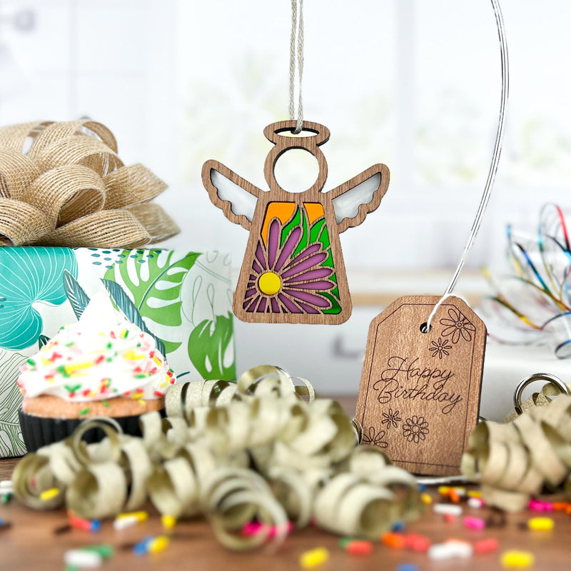 The Forged Flare® Mother's Angels® Aster Ornament—a 3.5" wooden angel with purple aster flower details—hangs by a “Happy Birthday” tag, surrounded by a gift box, cupcake with sprinkles, curly ribbons, and a burlap bow.