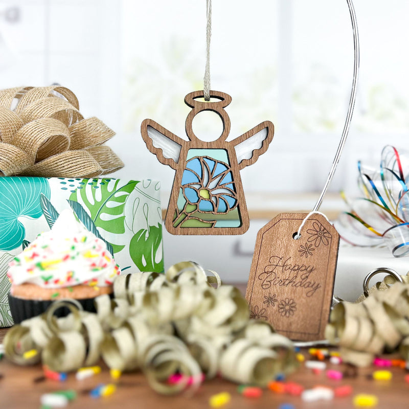 A Forged Flare® Mother's Angels® Morning Glory Ornament (3.5" angel figurine) hangs by a "Happy Birthday" tag near a sprinkle cupcake, wrapped gift, ribbon curls, and confetti on the table.
