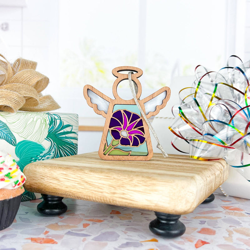 The Forged Flare® Purple Morning Glory Ornament, a 3.5" Mother's Angels® angel figurine with colorful stained glass, stands on a small platform in a bright room, surrounded by wrapped gifts, a cupcake, and a wire decoration.
