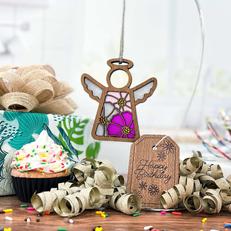 A Forged Flare® Mother's Angels® Cosmos Ornament (3.5" Angel Figurine) with pink and purple cosmos flowers hangs over a table set with a birthday cupcake, wrapped gift, gold ribbon curls, and a "Happy Birthday" wooden tag.