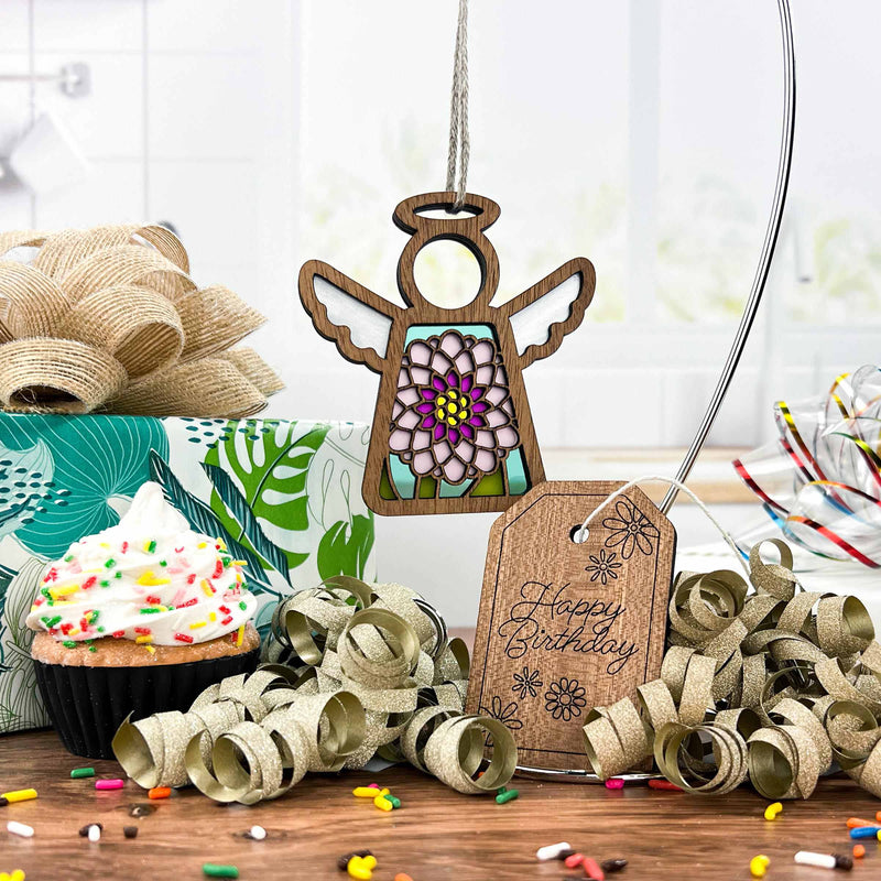 A Forged Flare® Mother's Angels® Chrysanthemum Ornament with imperfections hangs near a sprinkled cupcake, golden ribbon curls, and a "Happy Birthday" tag on the kitchen counter.