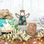 A Forged Flare® Mother's Angels® 3.5" Holly Ornament angel figurine hangs with a “Happy Birthday” tag, curly ribbons, holly flowers, a sprinkled cupcake, and wrapped gifts for a festive celebration scene.