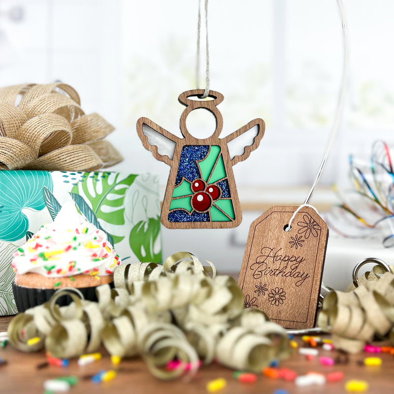 A Forged Flare® Mother's Angels® 3.5" Holly Ornament angel figurine hangs with a “Happy Birthday” tag, curly ribbons, holly flowers, a sprinkled cupcake, and wrapped gifts for a festive celebration scene.