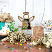 A Forged Flare® Mother's Angels® 3.5" stained glass-style wooden angel and Narcissus Ornament, December's birth flower, rest with a sprinkle cupcake, gifts, gold ribbons, and a “Happy Birthday” tag on a sunny kitchen table.