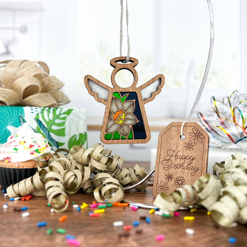 A Forged Flare® Mother's Angels® 3.5" stained glass-style wooden angel and Narcissus Ornament, December's birth flower, rest with a sprinkle cupcake, gifts, gold ribbons, and a “Happy Birthday” tag on a sunny kitchen table.