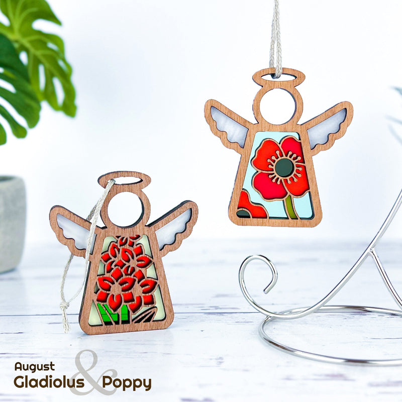 Two Forged Flare® Mother's Angels® 3.5" angel figurine ornaments—one with a red gladiolus and one with a red poppy—are shown on a white surface, text reading "August Gladiolus & Poppy," with green plants in the background.