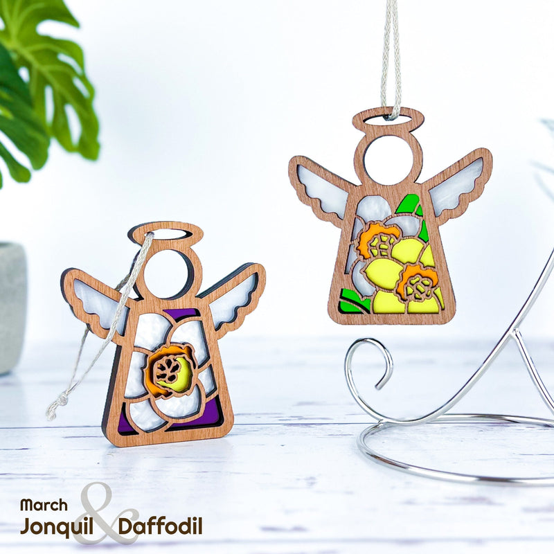 The Forged Flare® Mother's Angels® Daffodil Ornament is a 3.5" wooden angel figurine with stained glass-style daffodils, perfect as a birth flower gift. Display includes one angel on a stand and another upright on a white surface with plants.