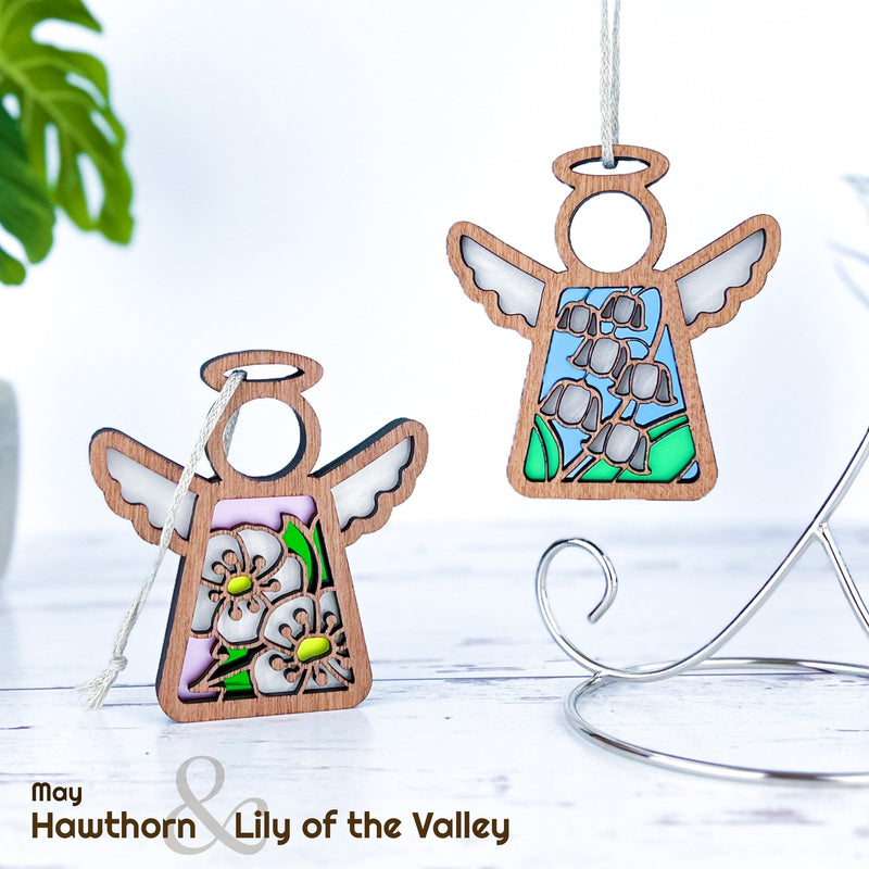 Two Forged Flare® Mother's Angels® wooden angel ornaments, 3.5" tall, feature stained glass-style Birth Flower designs: one with pink Hawthorn, the other with blue Lily of the Valley. Text: "may Hawthorn & Lily of the Valley.
