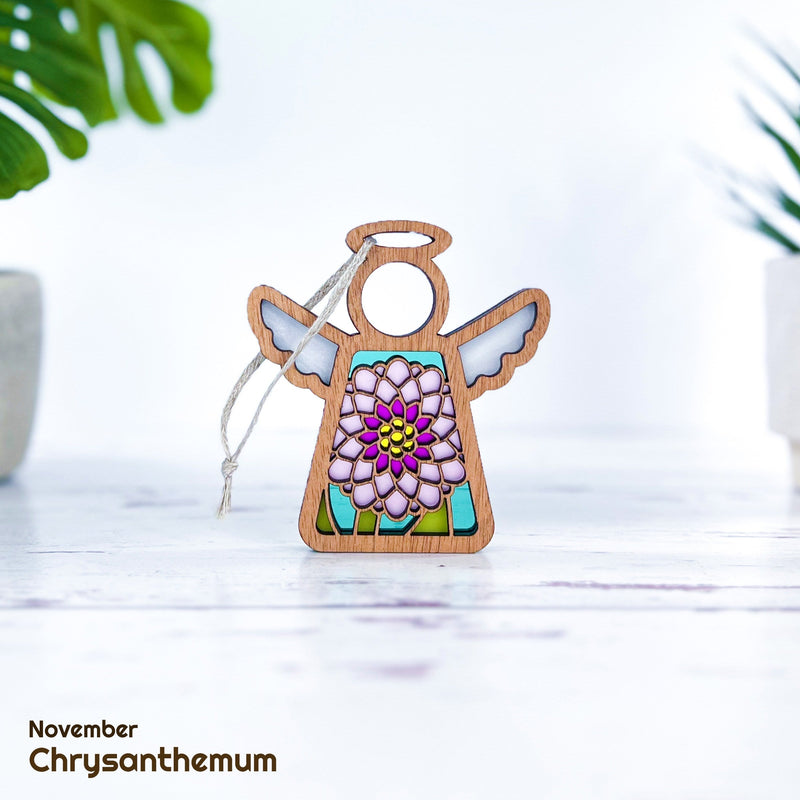 A Forged Flare® Mother's Angels® Chrysanthemum Ornament with Imperfections, a 3.5" handcrafted wooden angel figurine, hangs by a string between two green potted plants. "November Chrysanthemum" text appears in the bottom left corner.