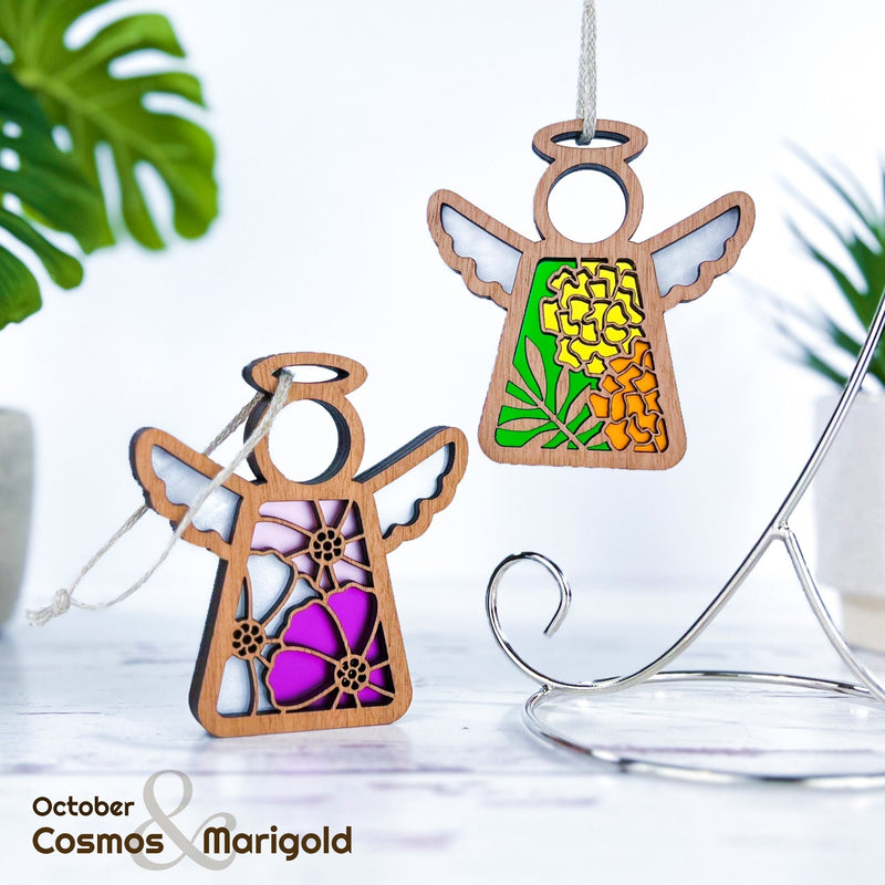 Two Forged Flare® Mother's Angels® ornaments, each 3.5" tall, display October birth flowers—cosmos and marigold. Both wooden angels hang by string among green leaves on a metal stand. Text reads "October Cosmos & Marigold.
