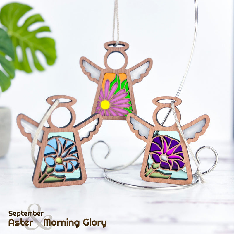 Three Forged Flare® Mother's Angels® 3.5" tall wooden angel ornaments feature stained glass-style aster and purple morning glory designs. Shown on a silver stand with greenery, text reads "September Aster & Morning Glory.
