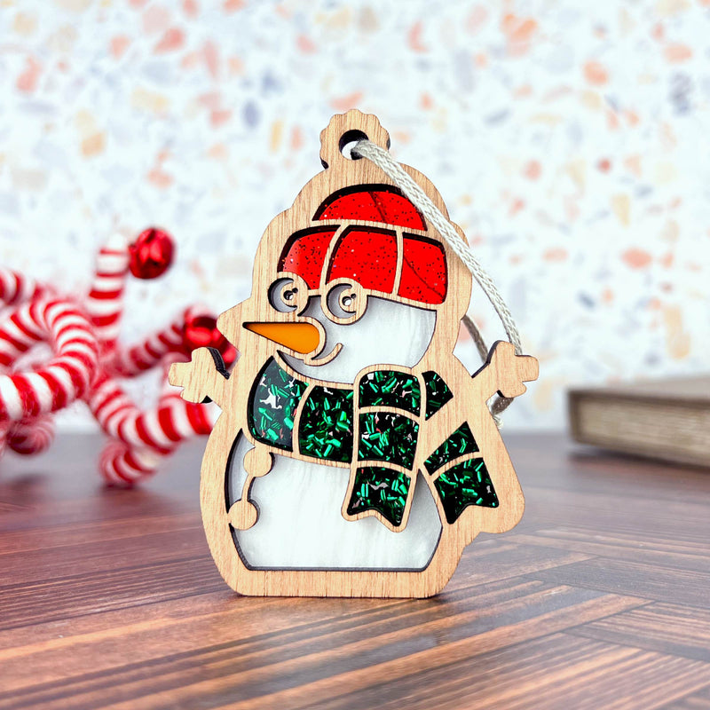 The Forged Flare® Christmas Snowman Ornament, 3.6" tall, features a wooden design with a red hat, green scarf, and glasses. It sits on wood surrounded by blurred peppermint candies and a closed book in the background.