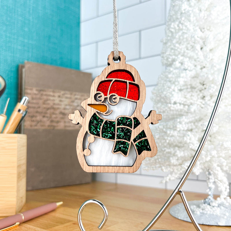 The Forged Flare® Christmas Snowman Ornament, 3.6" (H), with a red hat, green scarf, and glasses hangs on a silver stand; a white tree, pencils, and turquoise wall appear in the background.