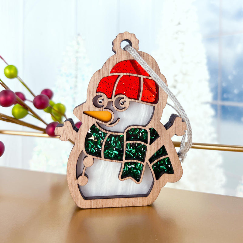 The Forged Flare® Christmas Snowman Ornament, 3.6" (H), features a red hat, green glittery scarf, and glasses. Set against blurred white trees and red berries, it adds a festive touch to any holiday décor.