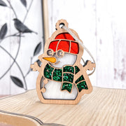 The Forged Flare® Christmas Snowman Ornament, 3.6" tall, features a red hat, green scarf, carrot nose, and hanging string. Ideal as a festive decoration or snowman gift, it stands on light wood with a bird silhouette in the background.