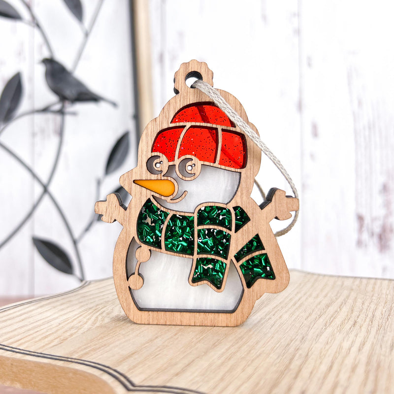 The Forged Flare® Christmas Snowman Ornament, 3.6" tall, features a red hat, green scarf, carrot nose, and hanging string. Ideal as a festive decoration or snowman gift, it stands on light wood with a bird silhouette in the background.