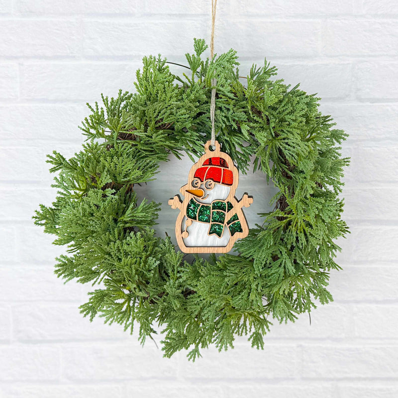 A Forged Flare® Christmas Snowman Ornament, 3.6" (H), with a red hat and green scarf, decorates a green holiday wreath on a white brick wall—an ideal festive accent for your home.