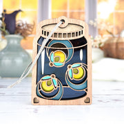 The Forged Flare® Fireflies Jar Ornament, 3.6" tall, features a mason jar design with three glowing blue and yellow fireflies and a hanging string. Soft autumn hues and vases are visible in the background through a window.