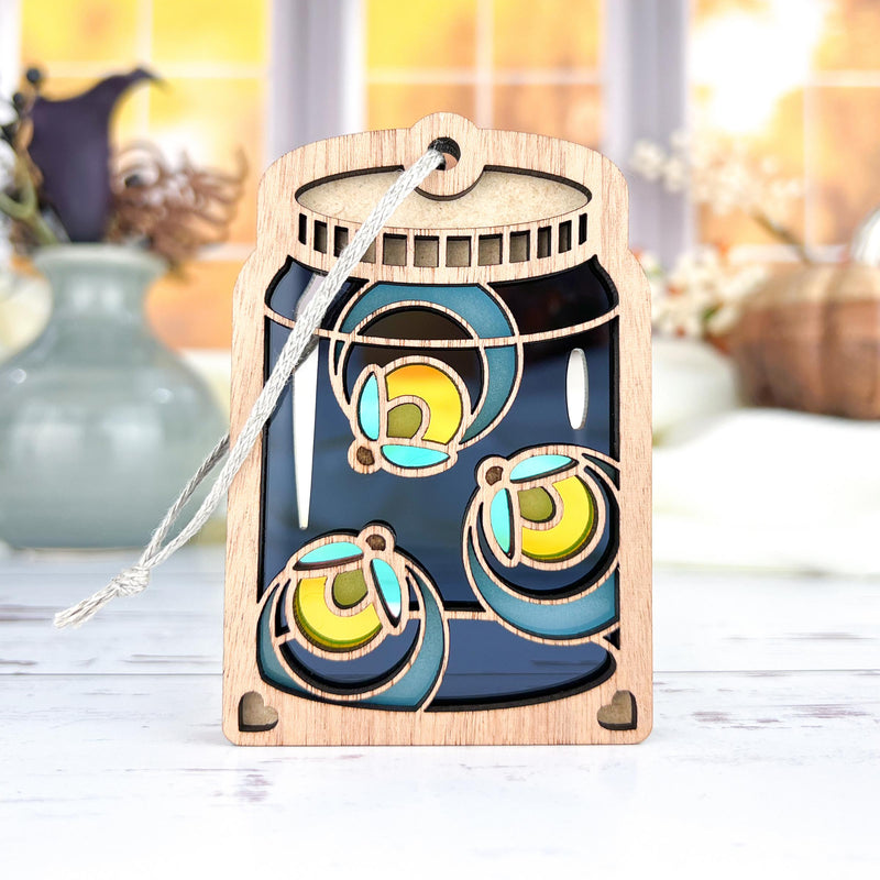 The Forged Flare® Fireflies Jar Ornament, 3.6" tall, features a mason jar design with three glowing blue and yellow fireflies and a hanging string. Soft autumn hues and vases are visible in the background through a window.