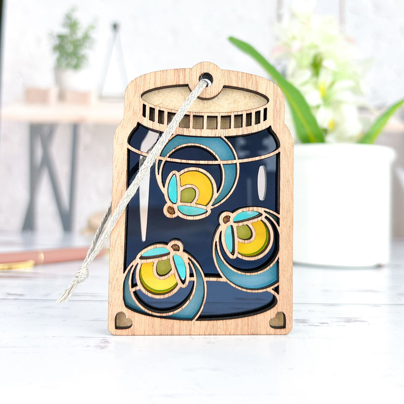 The Forged Flare® Fireflies Jar Ornament, 3.6" tall, is a decorative wooden tag shaped like a mason jar with a layered fireflies design on a dark background and includes a string loop for hanging. Background items appear blurred.
