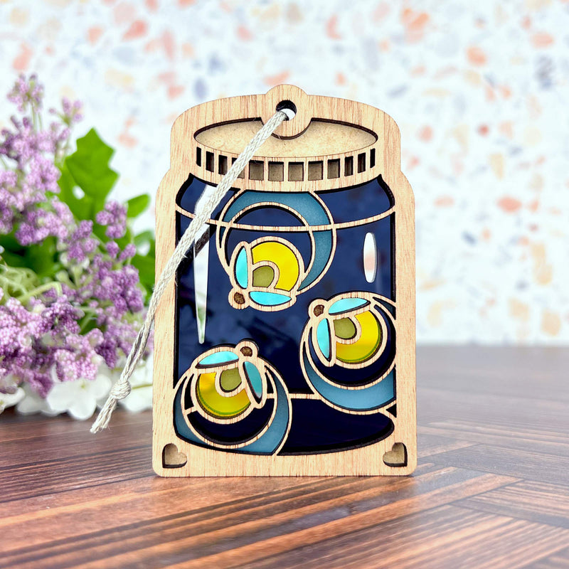 The Forged Flare® Fireflies Jar Ornament, 3.6" (H), is a wooden Christmas ornament featuring colorful citrus slices and glow in the dark charm, hanging by a silver string—perfect for festive décor.