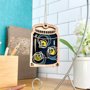 The Forged Flare® Fireflies Jar Ornament, 3.6" (H), hangs by a string and adds festive charm to any desk, displayed with pens, a notebook, and white flowers in the background.