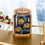 The Forged Flare® Fireflies Jar Ornament, 3.6" (H), is a wooden Christmas decoration with blue and green marbles, cutout details, a hanging string, and a festive glow in the dark effect against winter scenery.